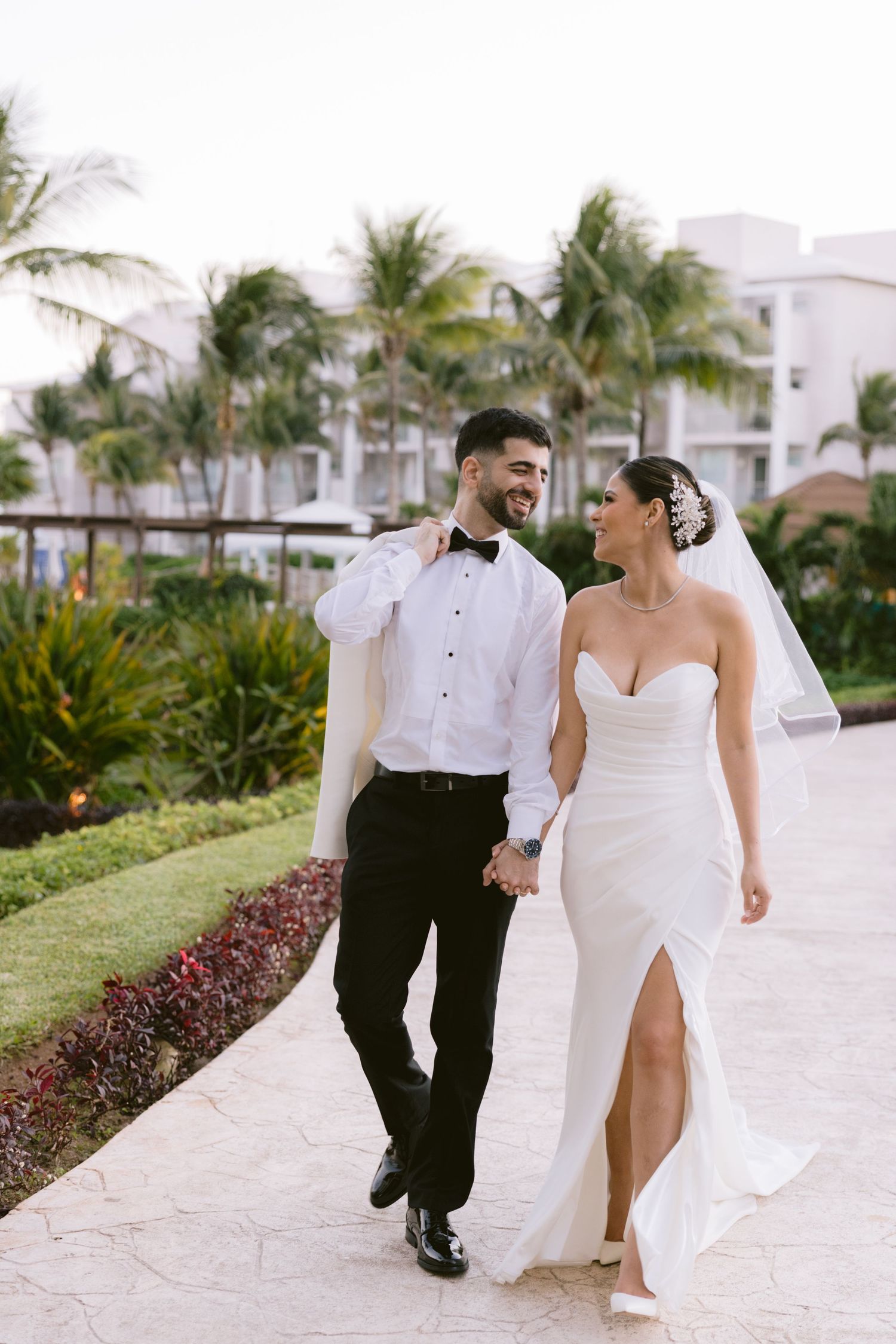 Bride and groom walk hand-in-hand at tropical resort wedding, groom in tuxedo, bride in strapless white gown with slit.