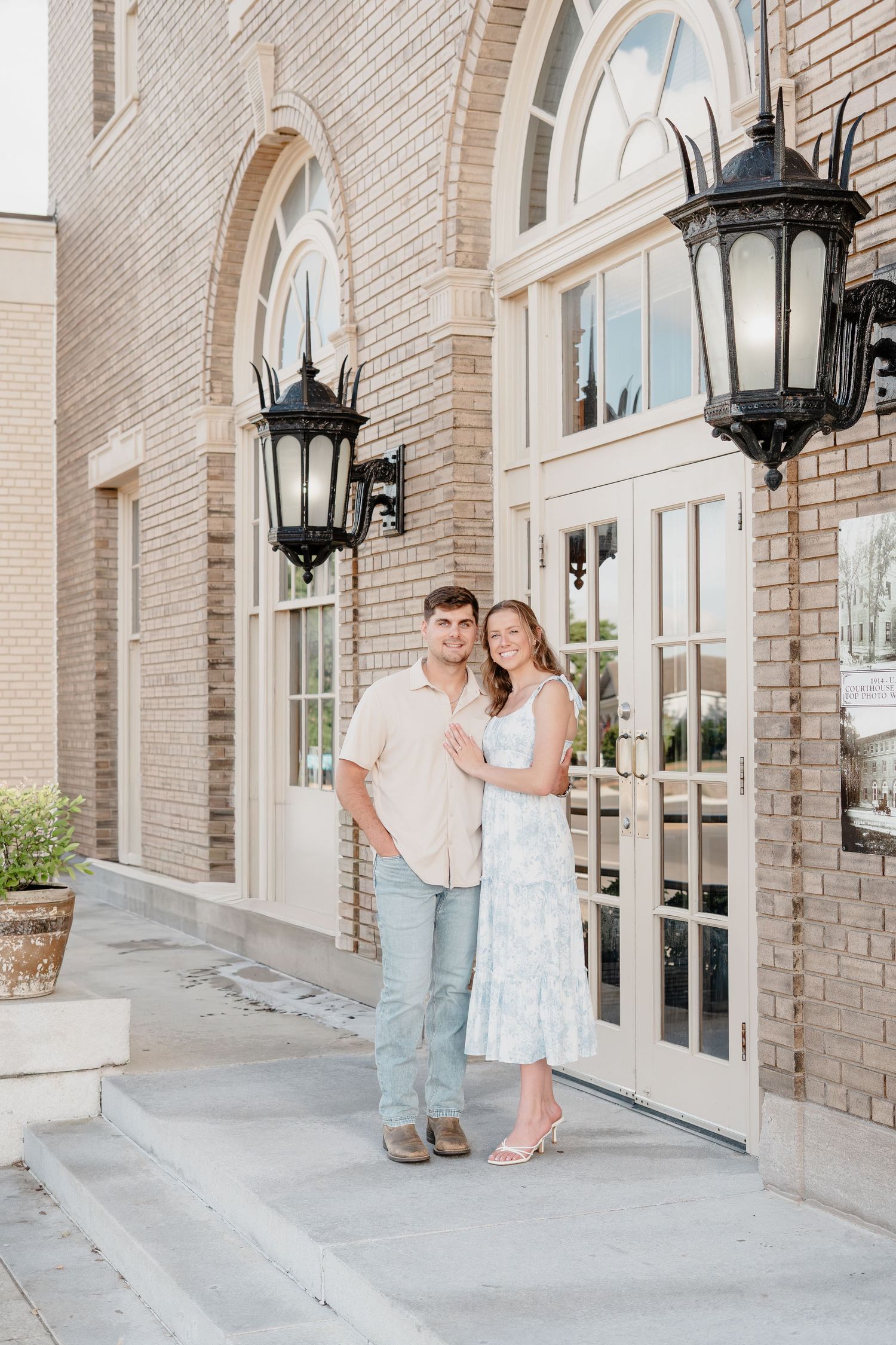 Couple posing together outside a brick building with elegant lanterns, man in pink shirt and woman in floral dress.