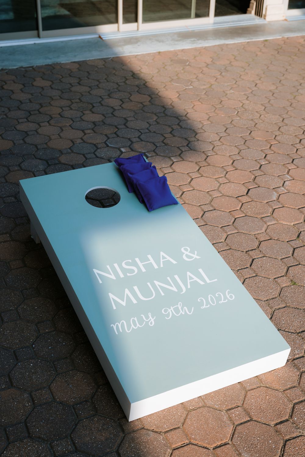 Custom cornhole board for Nisha and Munjal wedding on May 9th, 2026, with blue bean bags on brick patio.