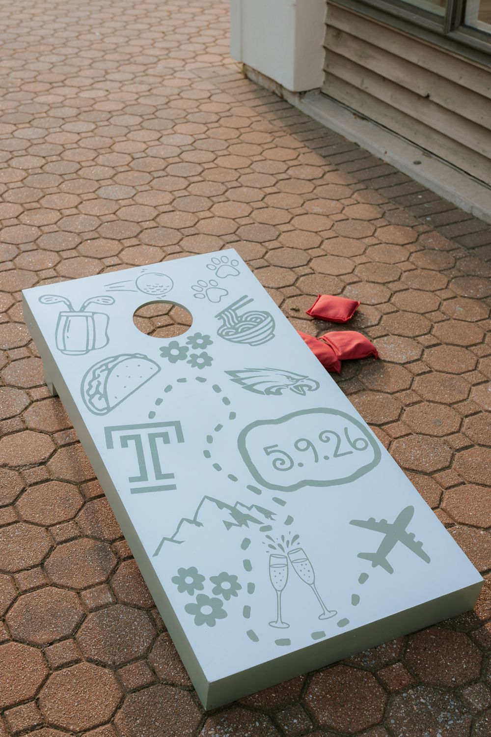 Custom cornhole board with wedding date 5.9.26, Temple University T logo, and travel icons on brick patio.