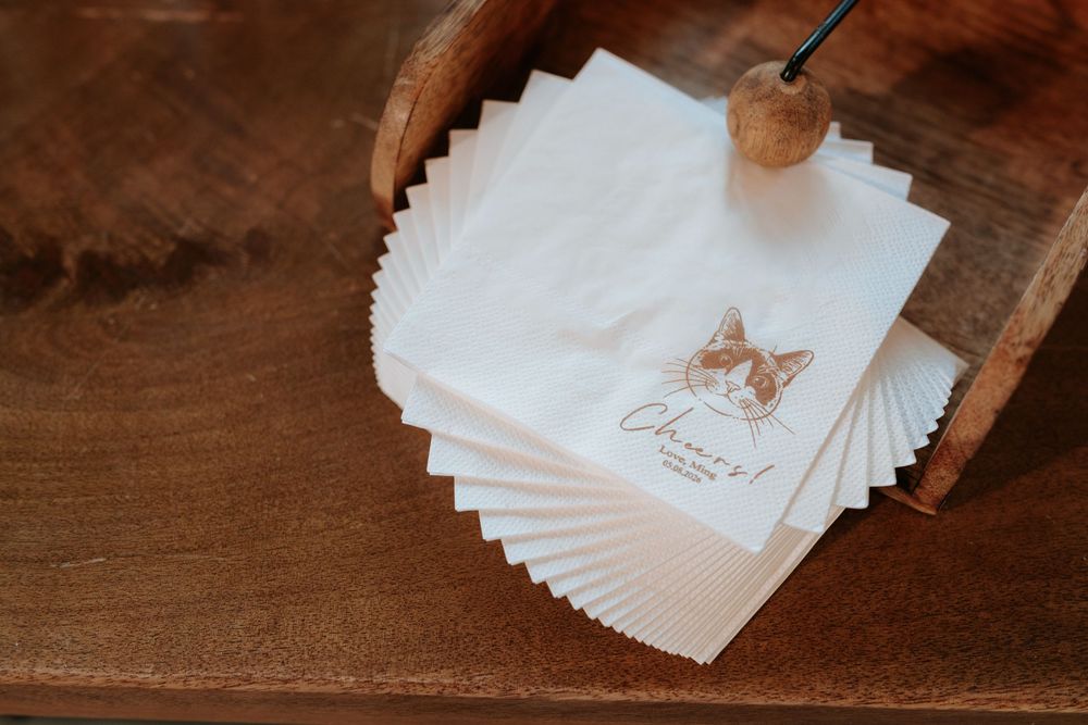 Custom printed white cocktail napkins featuring a cat illustration and "Cheers!" text, displayed in a wooden holder.