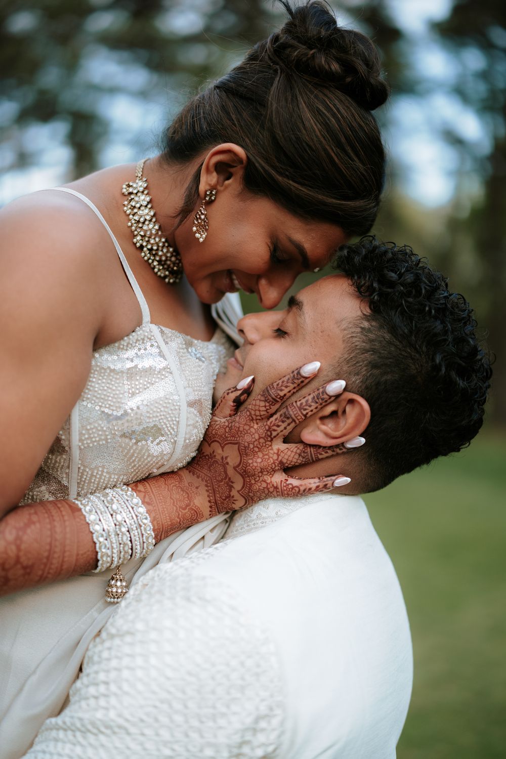 South Asian bride with henna hands and bangles embraces groom in white, both smiling during romantic wedding portrait.