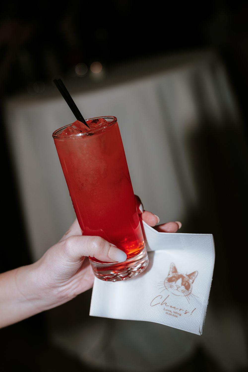 Hand holding a red cocktail with black straw and a cat-themed napkin reading Cheers at a bar event.