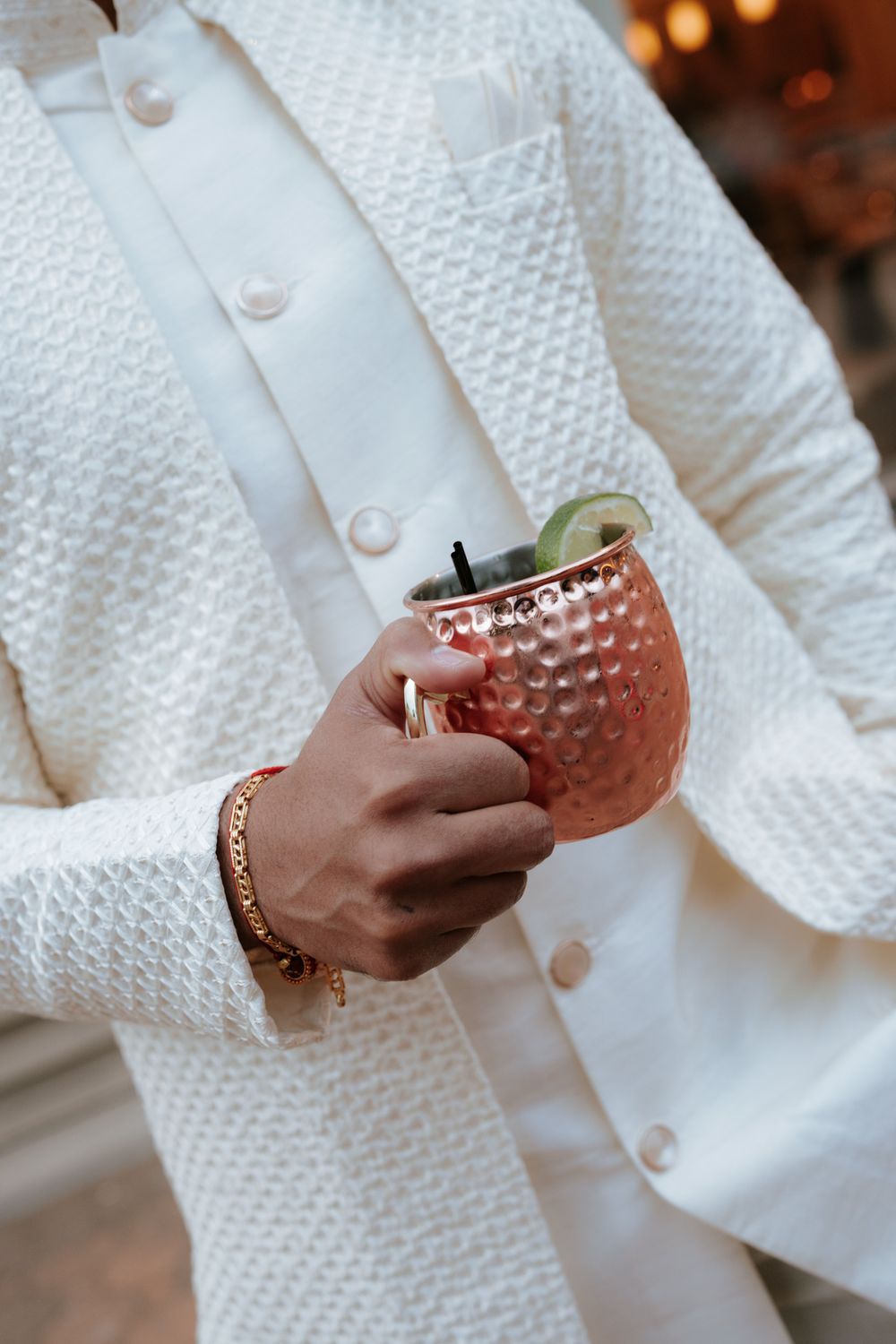Person in white textured suit holding a copper Moscow Mule mug with lime garnish and gold bracelet.