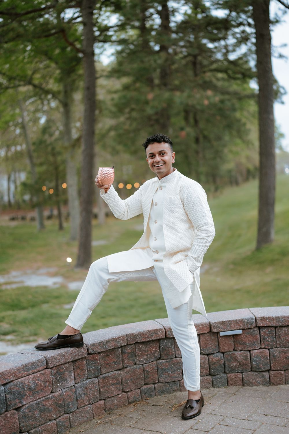A smiling man in a white Indian kurta suit holds a drink while posing on a stone wall outdoors.
