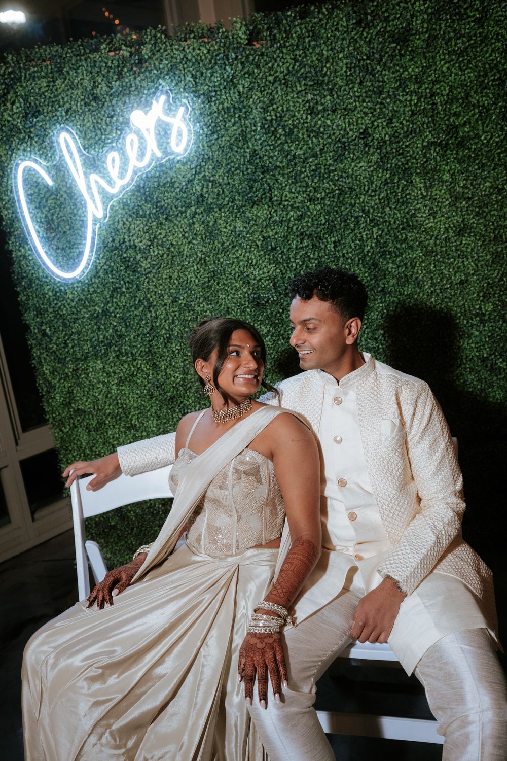 Couple in white Indian wedding attire sitting before a green hedge wall with a glowing Cheers neon sign.