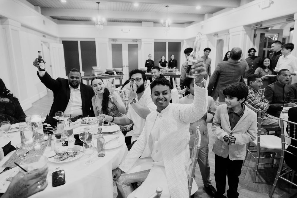 Black and white photo of joyful guests raising glasses in a toast at an elegant indoor wedding reception celebration.
