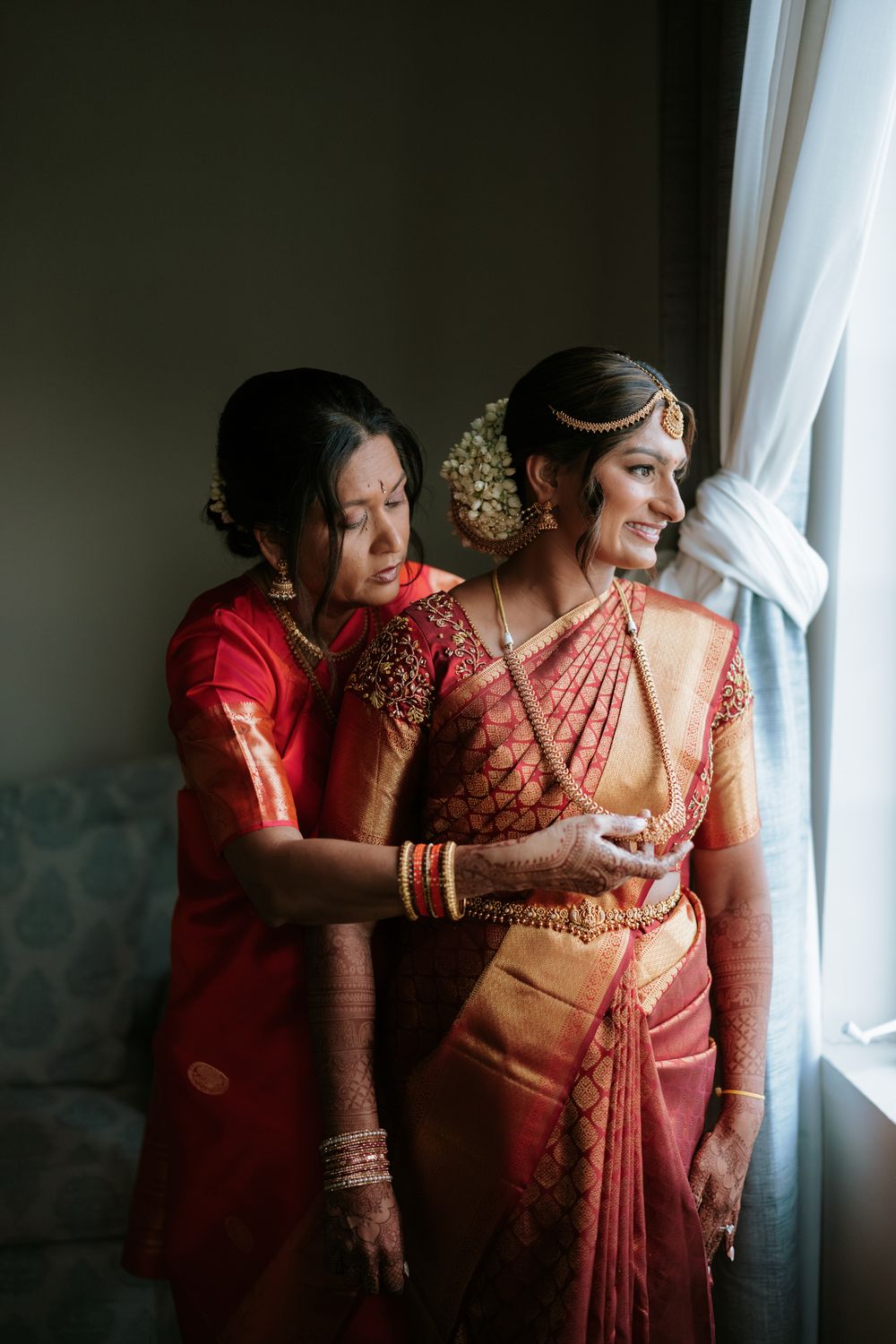 Two women in traditional red and gold Indian sarees stand by a window, one helping the bride prepare for her wedding ceremony.