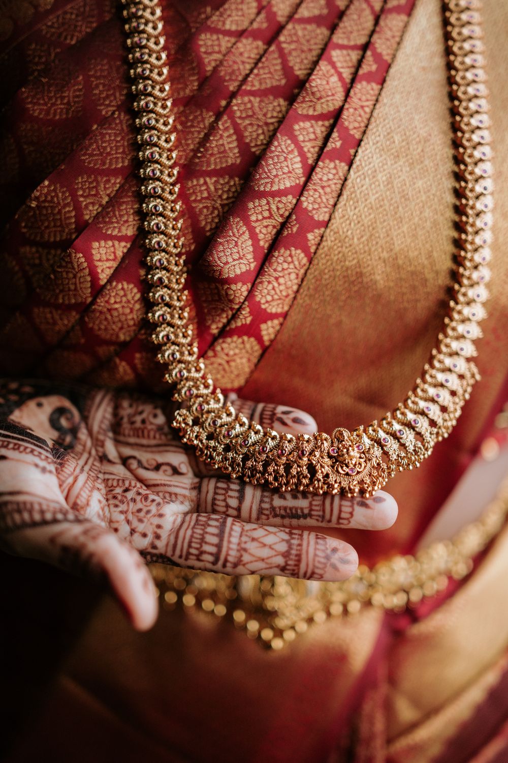 Close-up of Indian bride holding gold temple jewelry necklace, adorned with intricate henna mehndi designs on hands, wearing red silk saree.