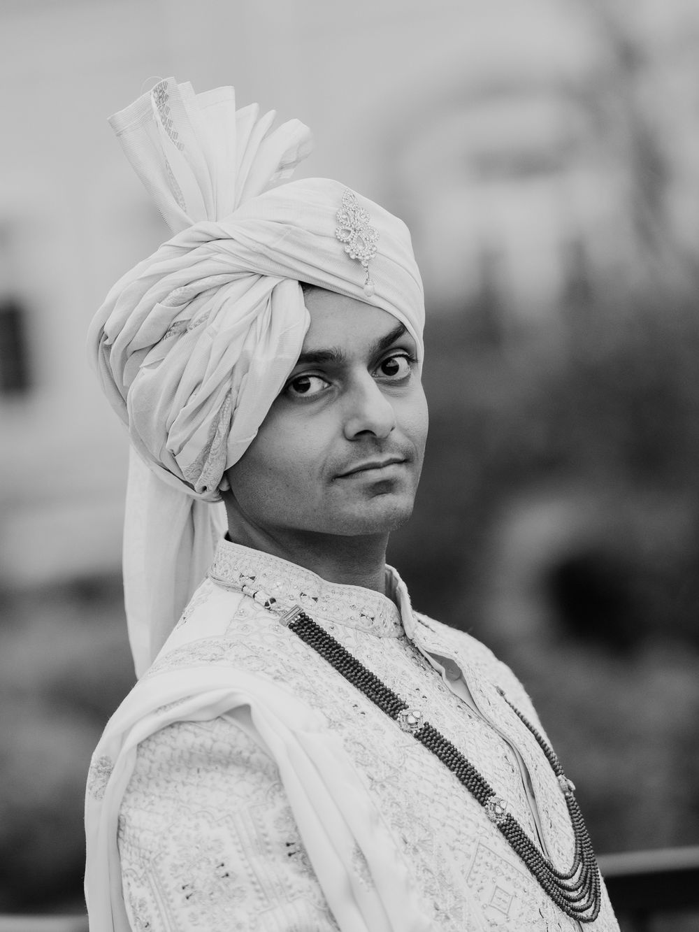 A groom in a white sherwani and turban poses elegantly in a black and white portrait photo.