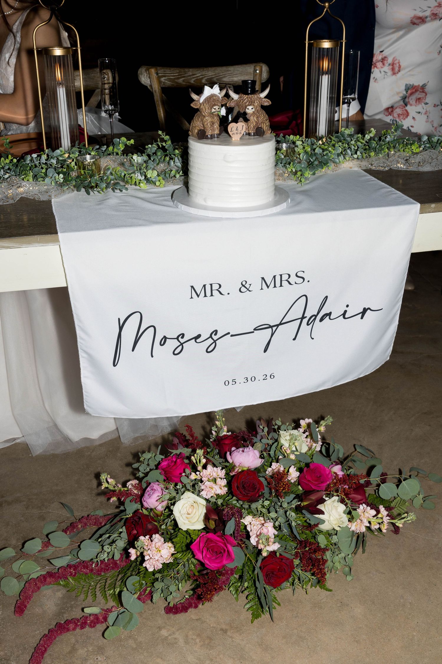 Wedding cake table with Mr. & Mrs. Moses-Adair sign, floral arrangement, and cow topper cake dated 05.30.26.