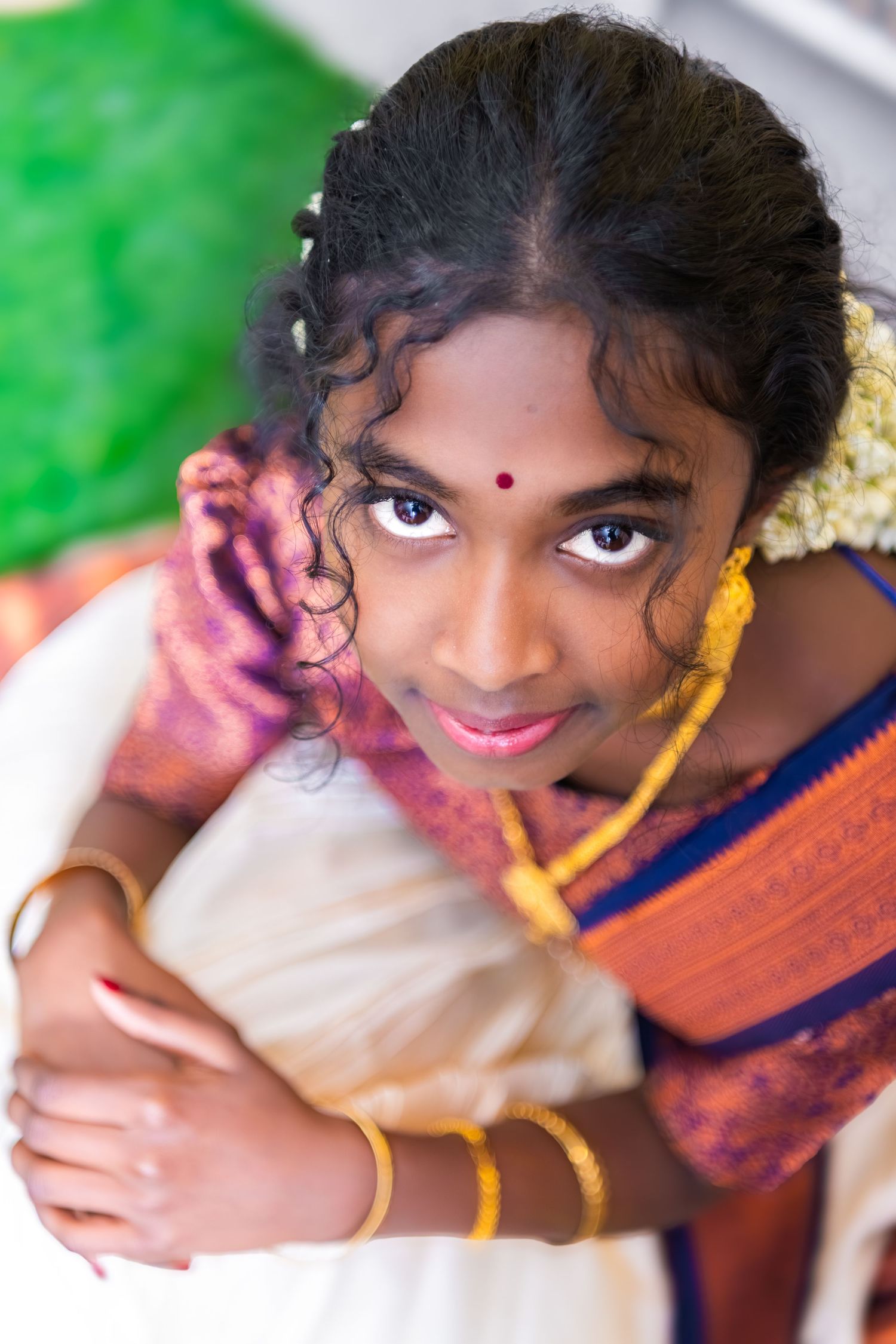 Young South Indian girl in traditional saree with gold bangles, bindi, and flower hair accessories smiling at camera.