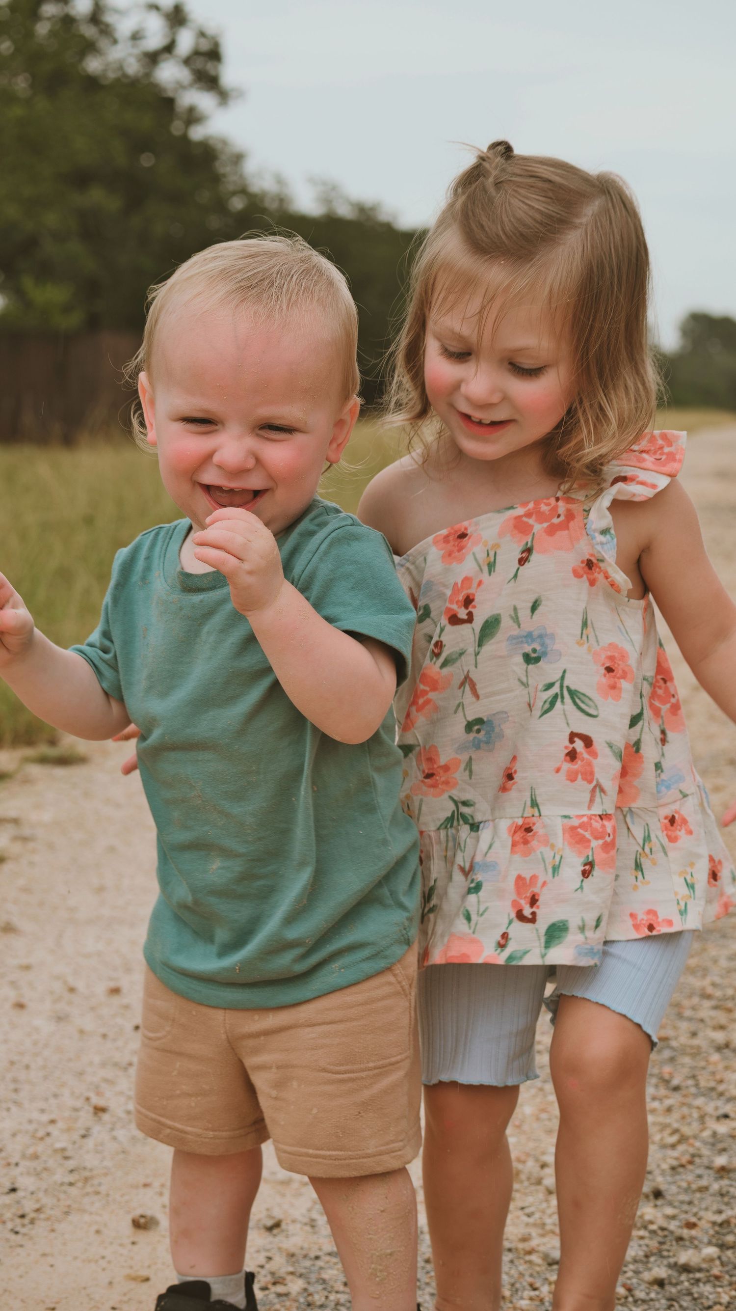 Two happy toddlers playing outdoors on a dirt path, boy in teal shirt and girl in floral dress.