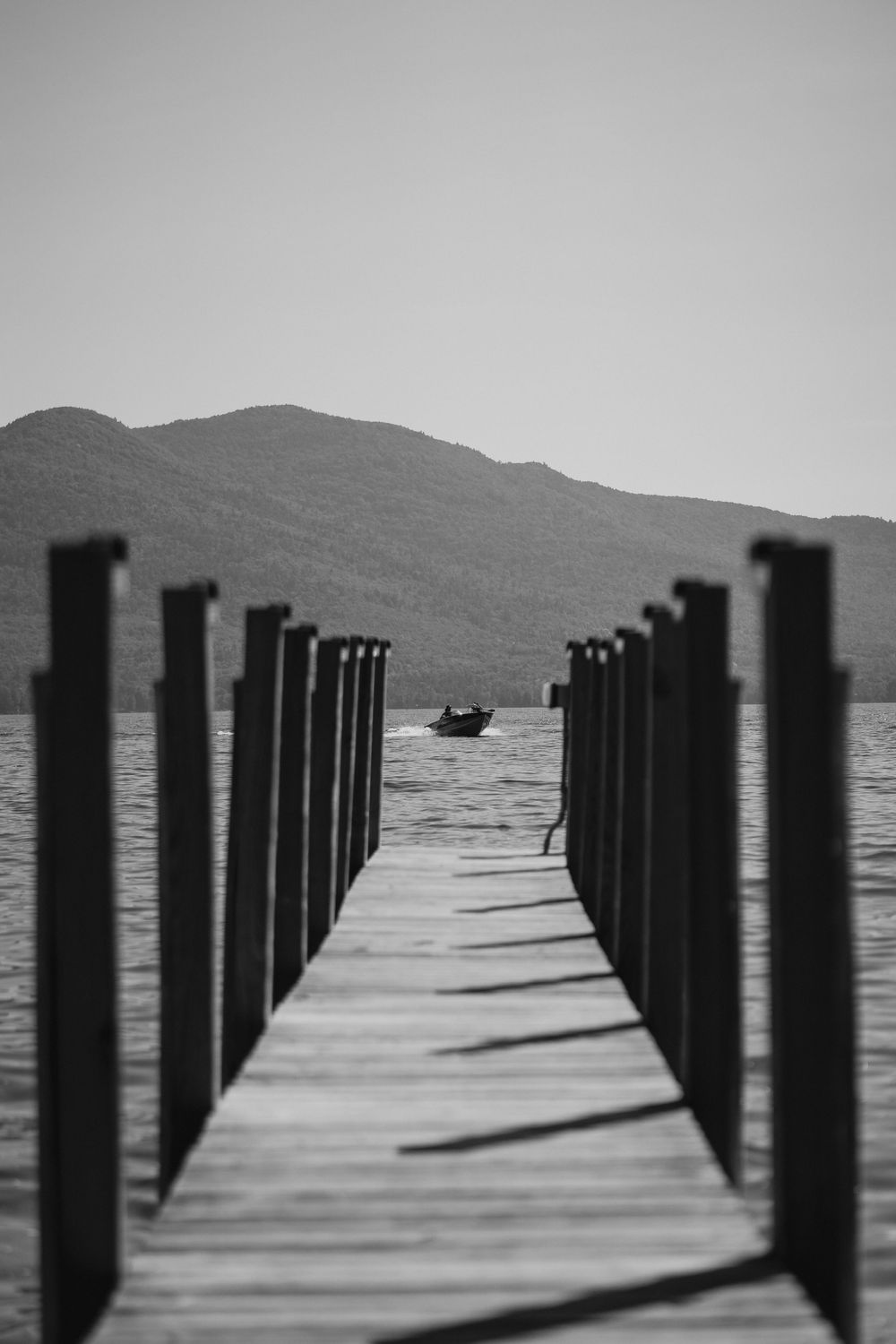 Black and white photo of a wooden dock leading to a calm lake with a boat and mountain backdrop.