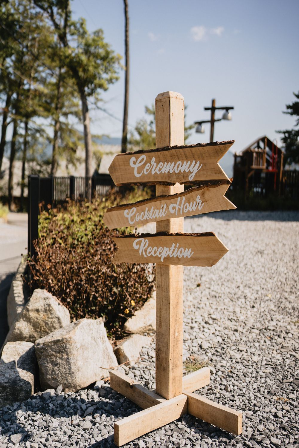 Rustic wooden wedding directional sign with arrows pointing to Ceremony, Cocktail Hour, and Reception on a gravel path.