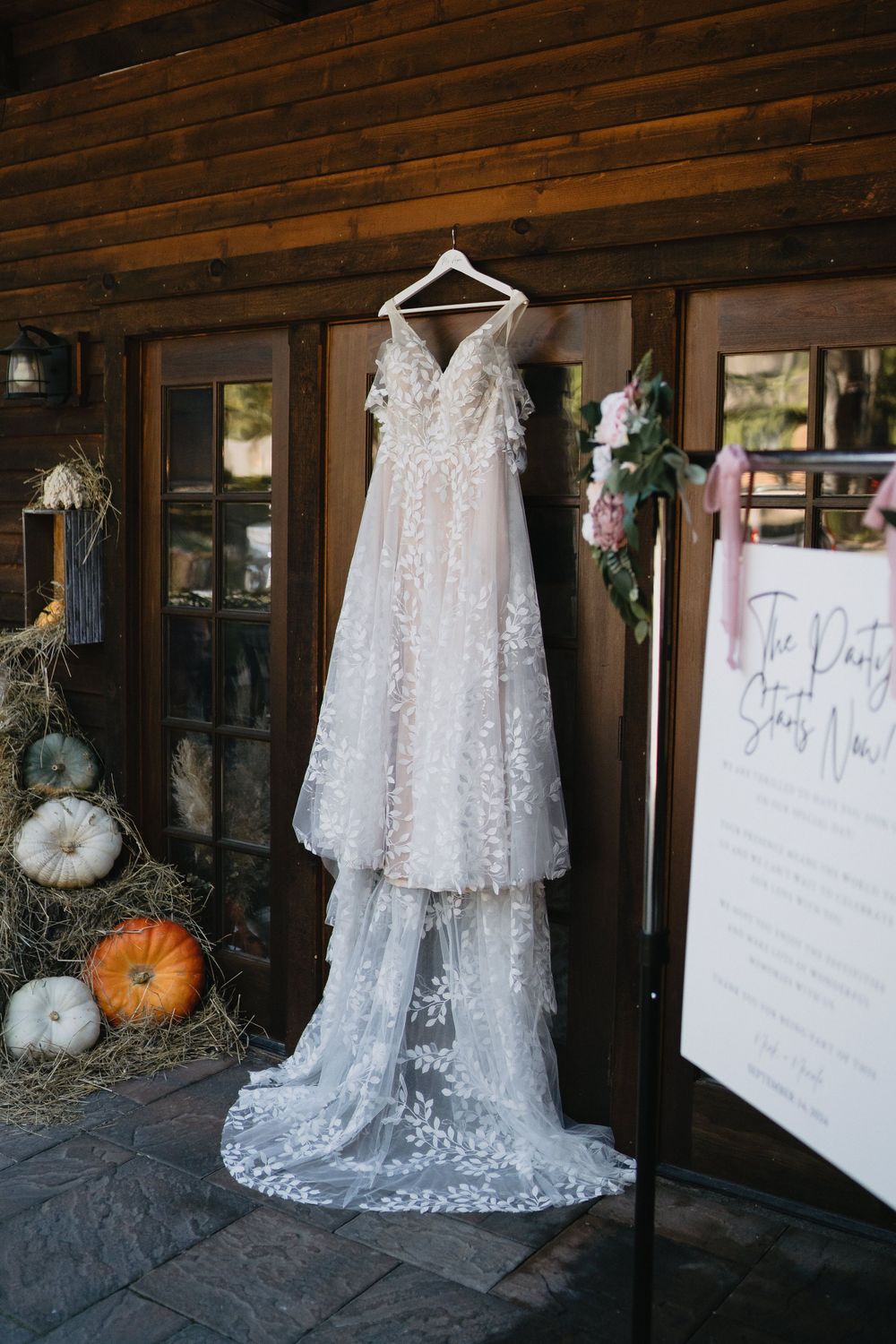 Elegant lace wedding gown with floral appliques hanging outside a rustic log cabin surrounded by fall pumpkin decorations.