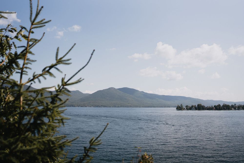 Serene lake view with a mountain backdrop, framed by pine tree branches under a partly cloudy blue sky.
