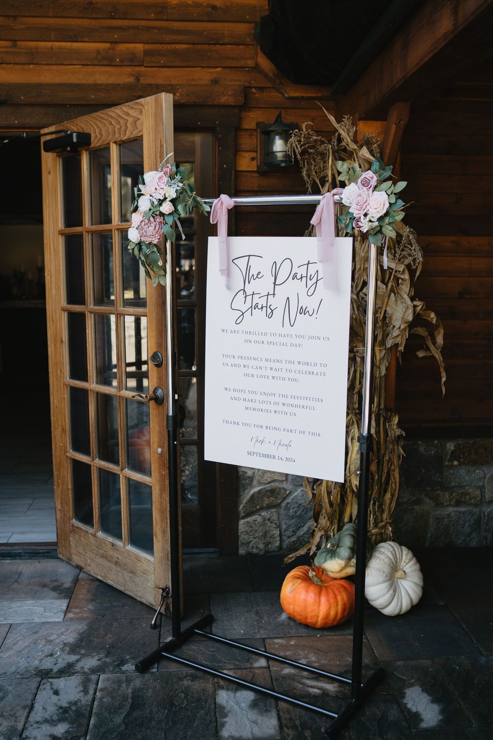 Rustic fall wedding welcome sign reading The Party Starts Now adorned with pink flowers and pumpkins at a log cabin venue.