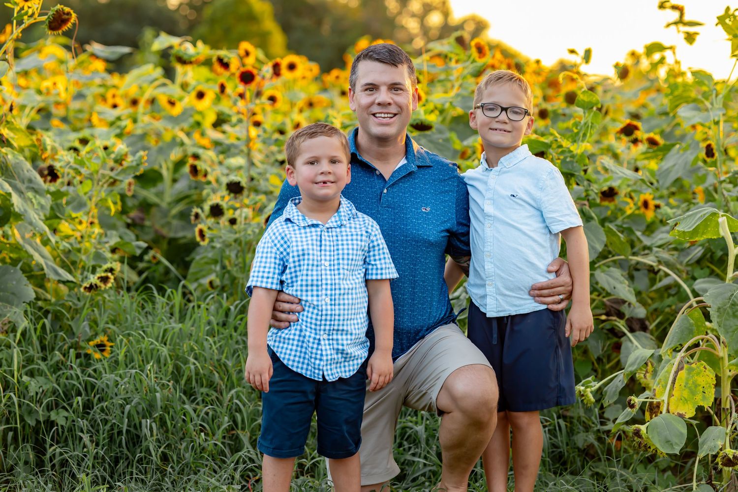 A man and two young boys smile in a sunflower field during golden hour.