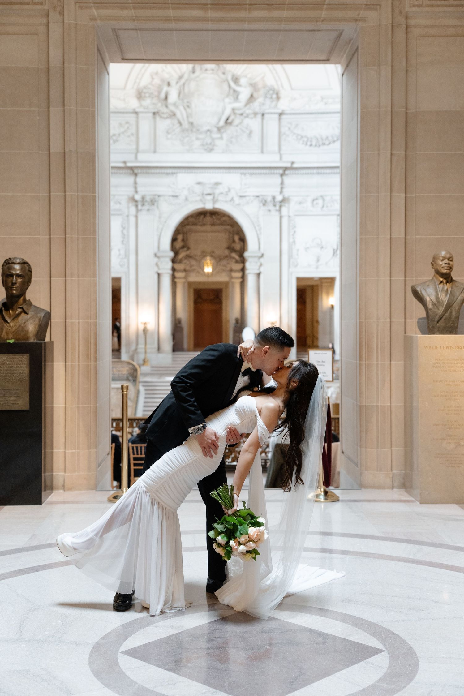 Groom dips bride for a romantic kiss inside San Francisco City Hall, her white gown and veil flowing gracefully.
