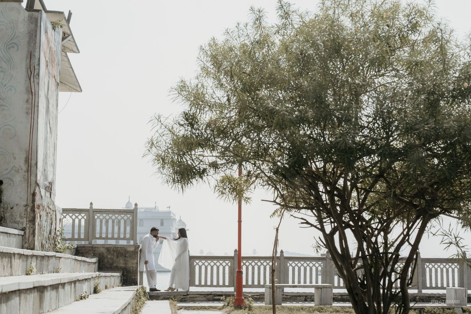 Couple in white attire standing on scenic terrace steps near a tree with misty coastal views.