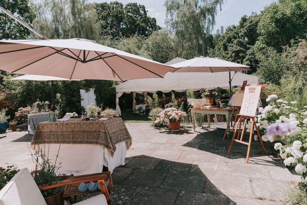 An outdoor wedding reception area with large pink umbrellas, floral arrangements, and tables on a sunny patio.