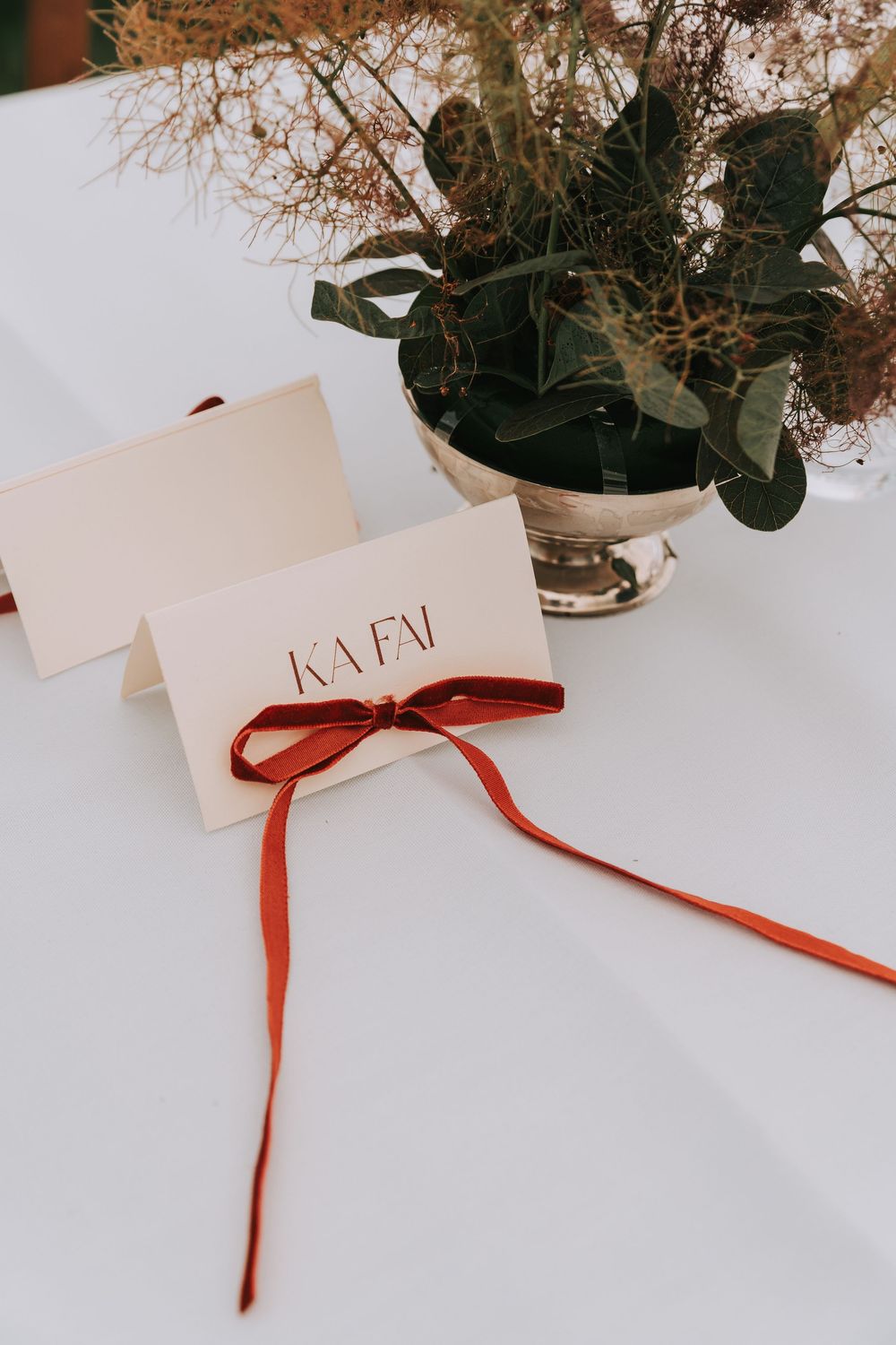 Wedding place card with red ribbon bow next to silver vase with eucalyptus and dried flowers on white tablecloth.