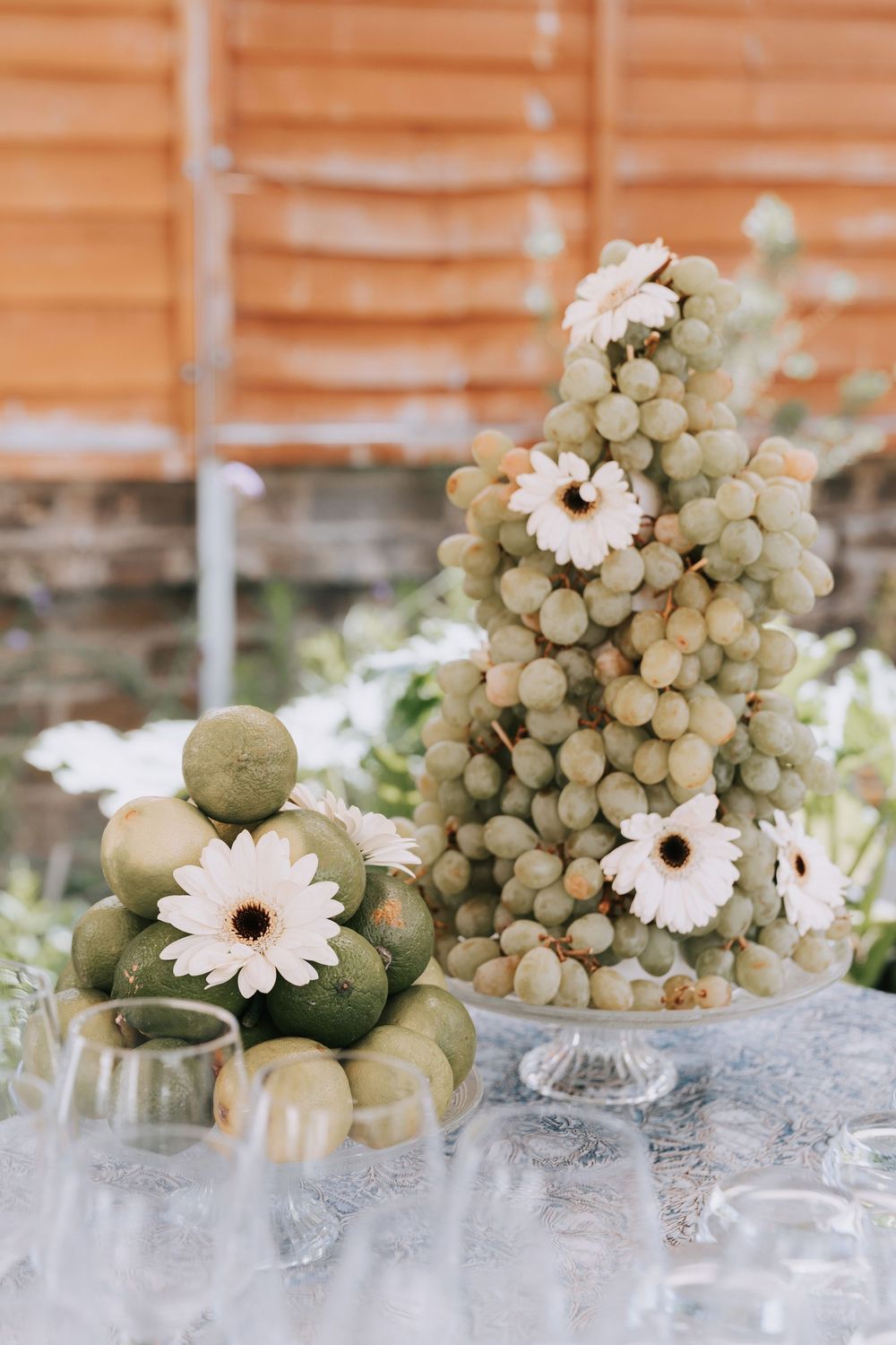 Elegant wedding dessert table with grape tower and green fruit pyramid decorated with white daisy flowers on lace tablecloth.