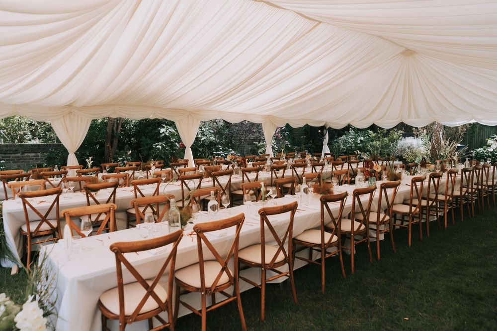 Elegant outdoor wedding reception under a white marquee tent with long wooden tables, cross-back chairs, and floral centerpieces.