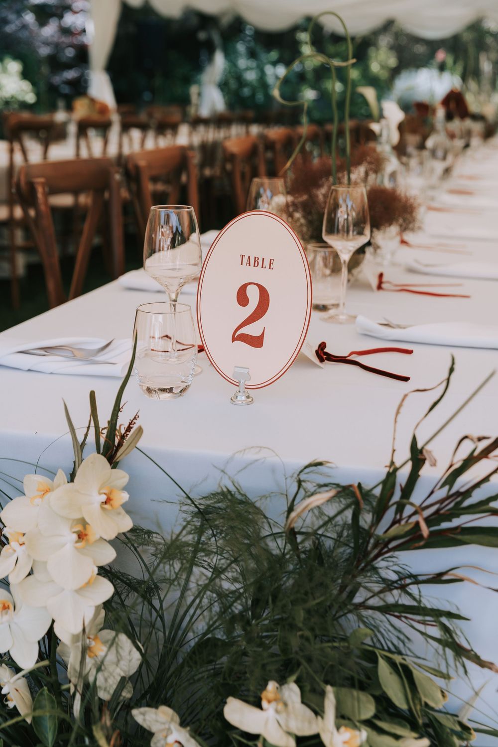 Elegant wedding reception table with white linen, floral centerpiece, wine glasses, and a red Table 2 sign.