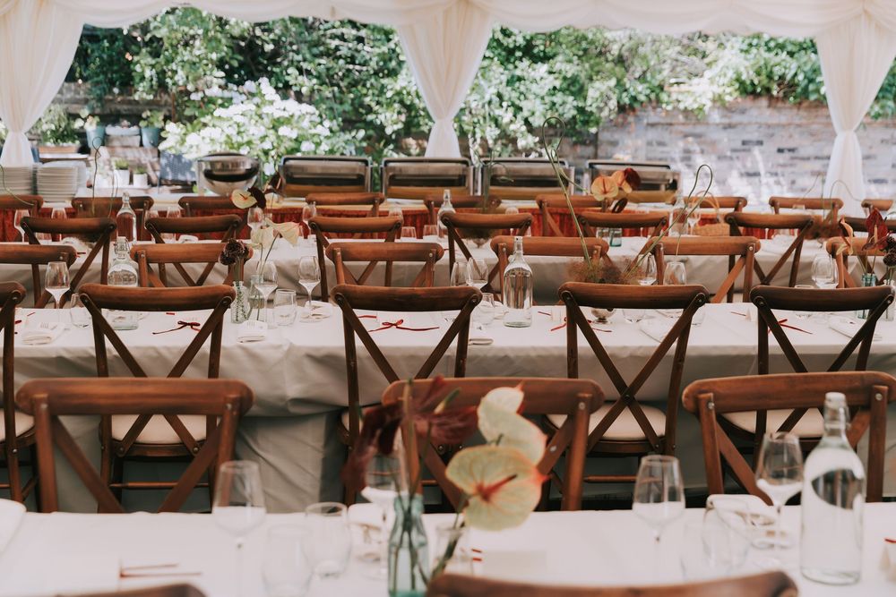 Elegant outdoor wedding reception tent with rustic wooden cross-back chairs and white linen tables adorned with floral centerpieces.