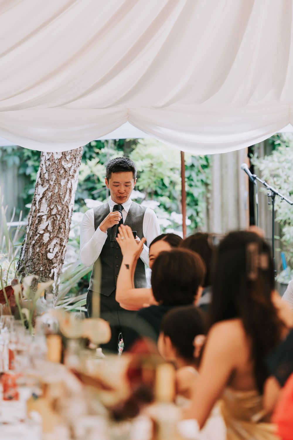 A man in a gray vest gives a speech at an elegant outdoor wedding reception under white draped fabric.
