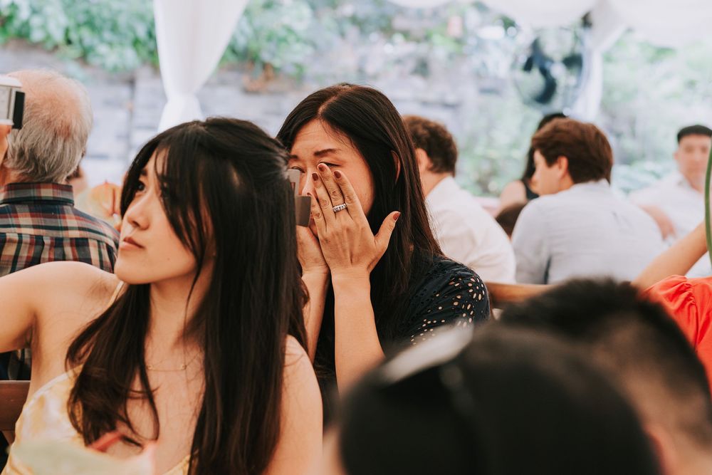 Emotional woman covering her mouth crying at a wedding reception seated next to another young woman.