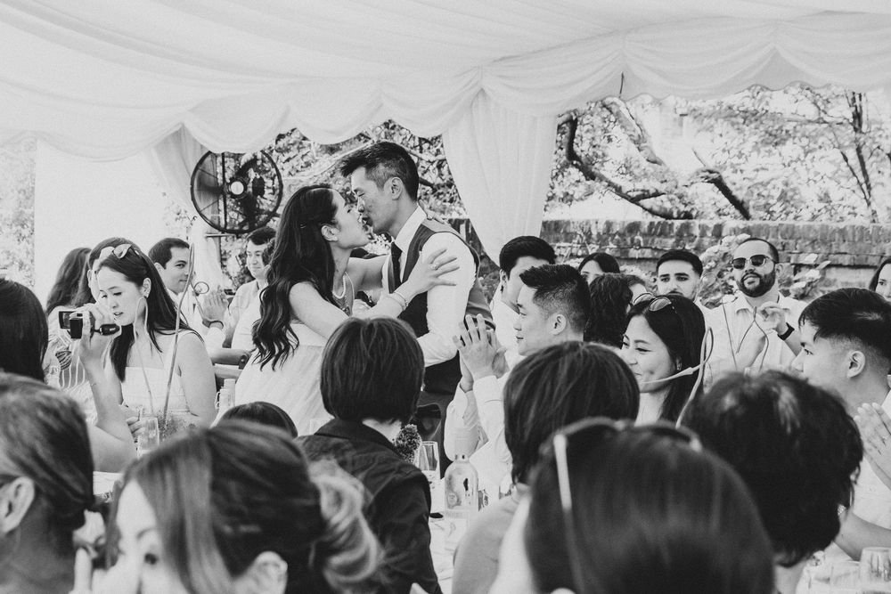 Black and white photo of a couple sharing a kiss at their wedding reception surrounded by smiling guests.