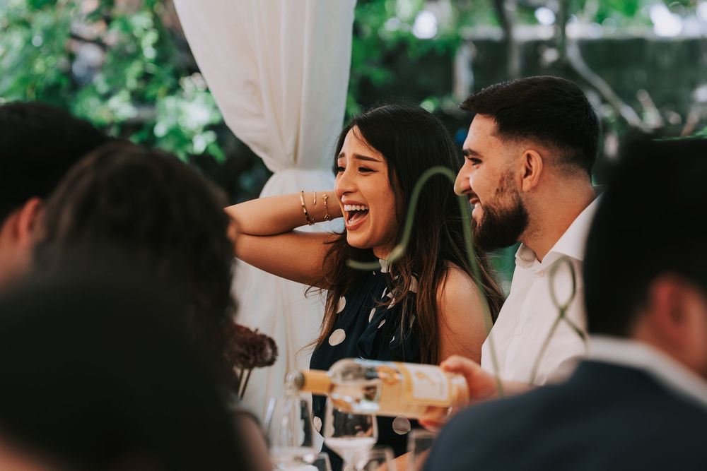 Guests laughing and enjoying conversation at an outdoor wedding reception dinner under a white tent.