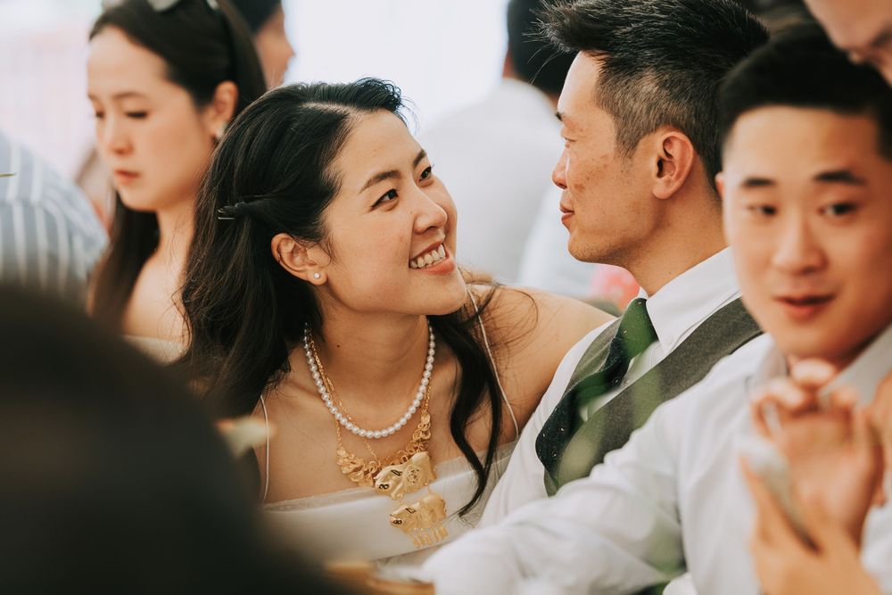 Joyful bride in pearl necklace laughing with groom in suit at wedding reception surrounded by guests.