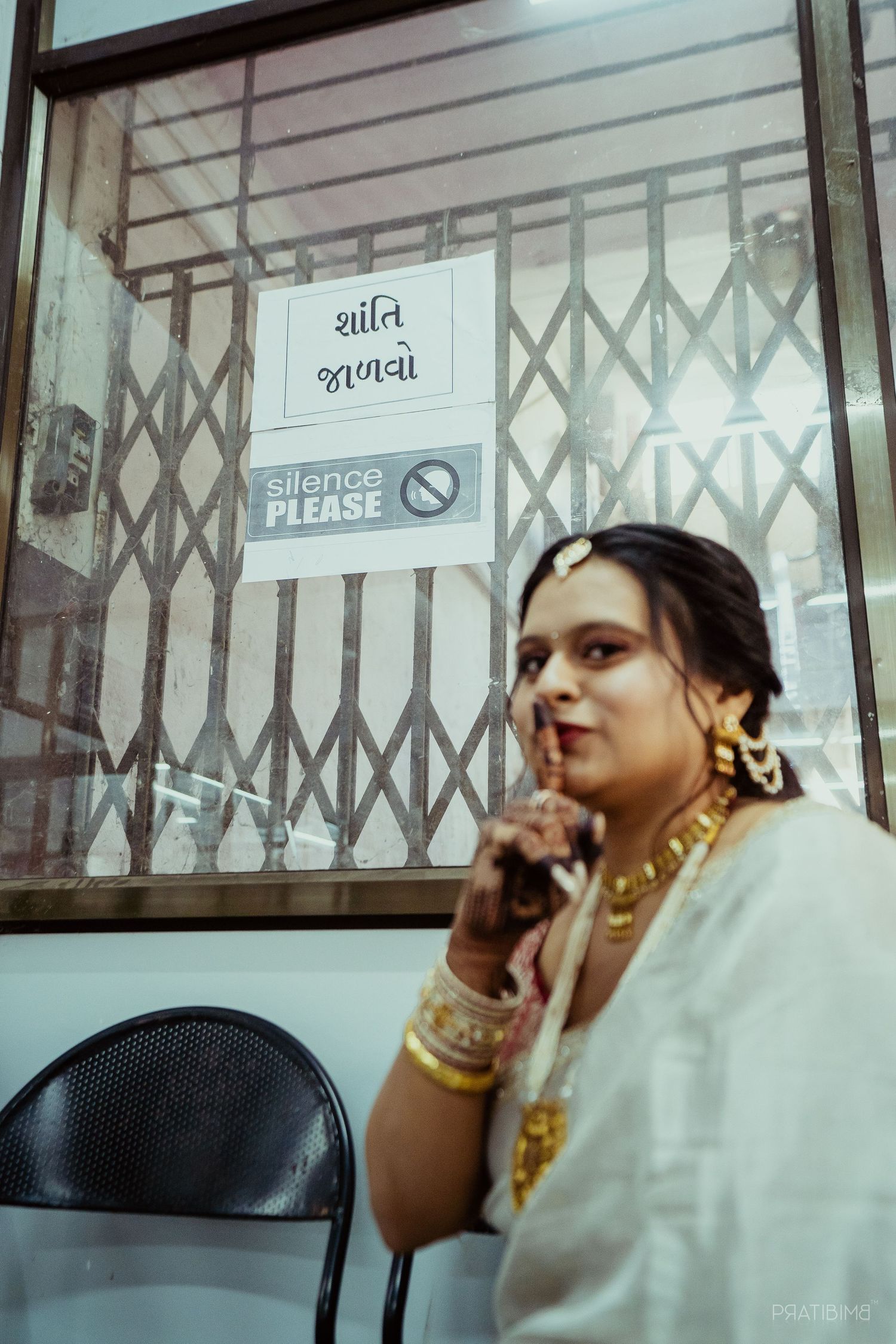 Indian bride in white saree with gold jewelry poses by a Silence Please sign, finger to lips.