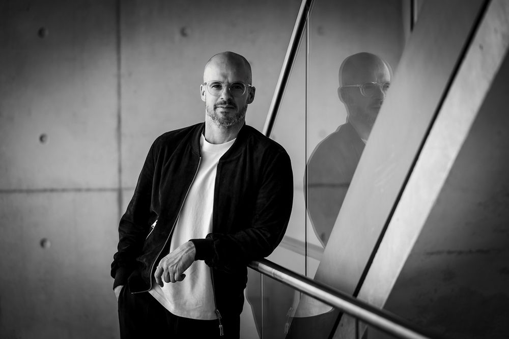 Schwarz-weißes Businessporträt eines Mannes, der sich an ein Treppengeländer lehnt und direkt in die Kamera blickt.