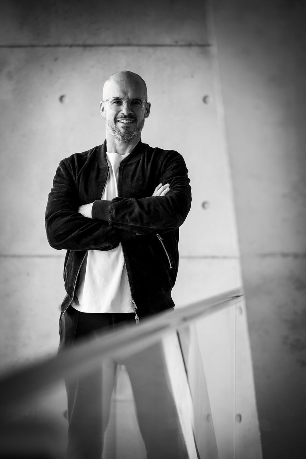 Schwarz-weißes Businessporträt eines lächelnden Mannes mit verschränkten Armen, weißem T-Shirt und dunklem Blouson.