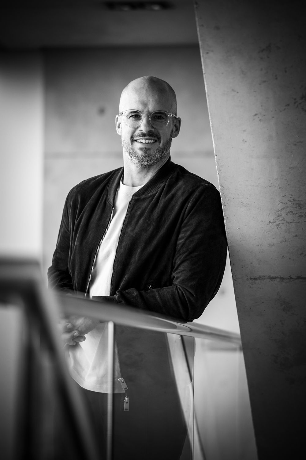 Schwarz-weißes Businessporträt eines Mannes mit Brille, der sich entspannt an ein Treppengeländer lehnt und in die Kamera lächelt.