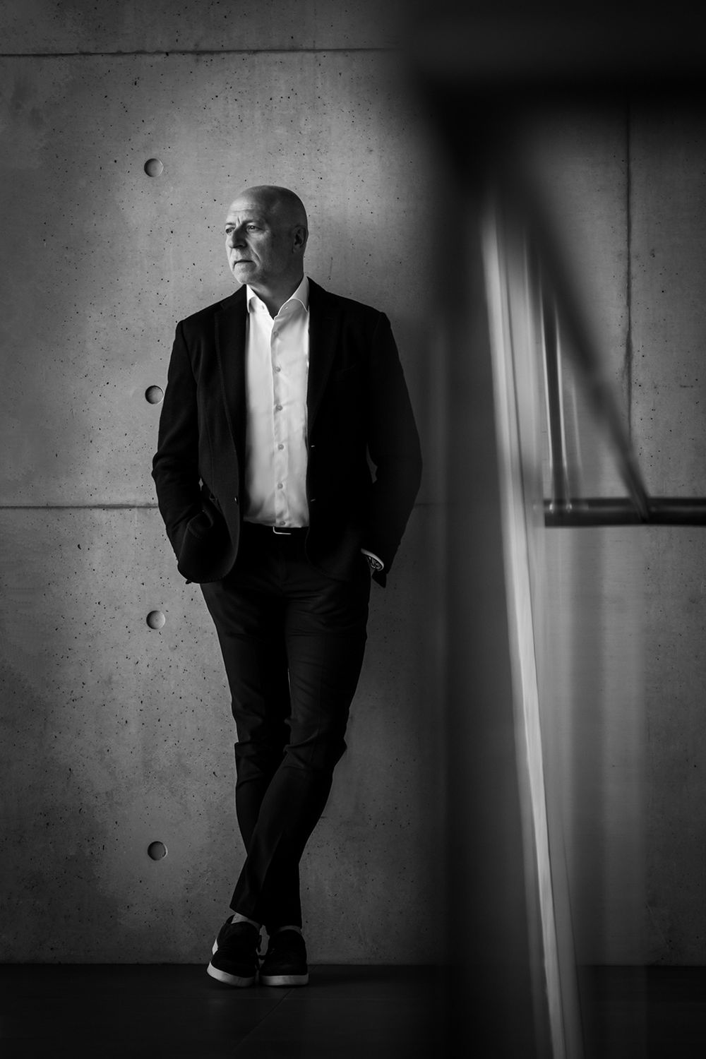 Businessporträt eines Mannes in Ganzaufnahme, an eine Betonwand gelehnt, mit Händen in den Hosentaschen und seitlichem Blick.