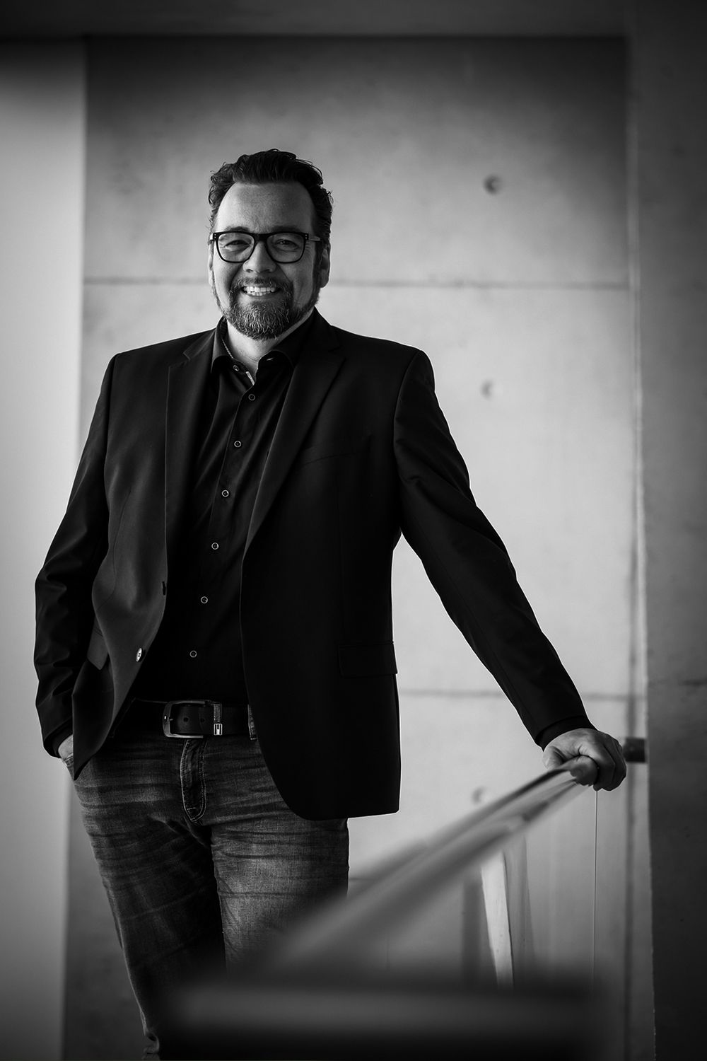 Businessporträt eines Mannes mit Brille, Vollbart und Jackett, der sich am Treppengeländer abstützt und in die Kamera lächelt.