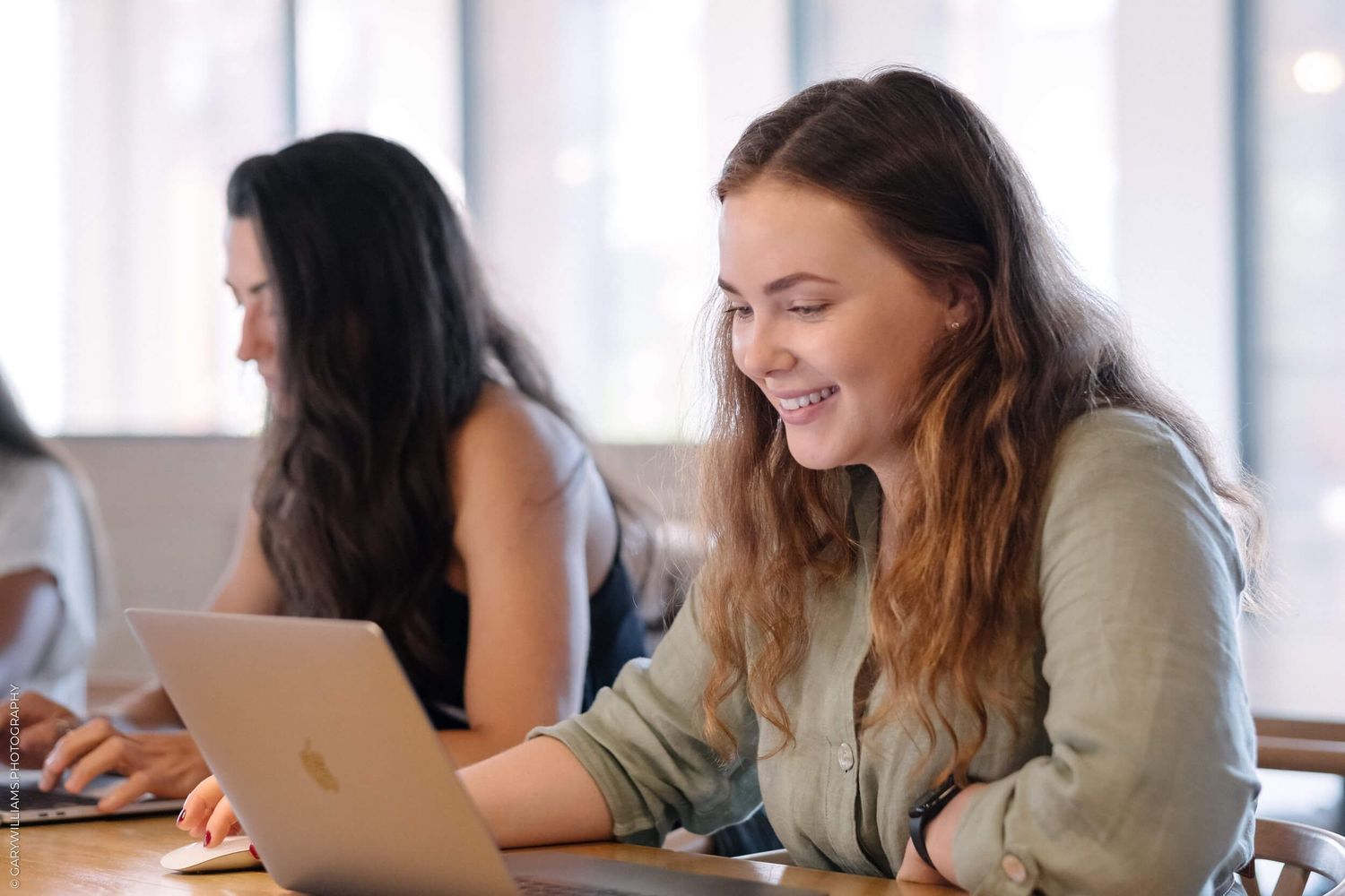 A smiling young woman works on a MacBook laptop at a desk, with another woman sitting in the background.