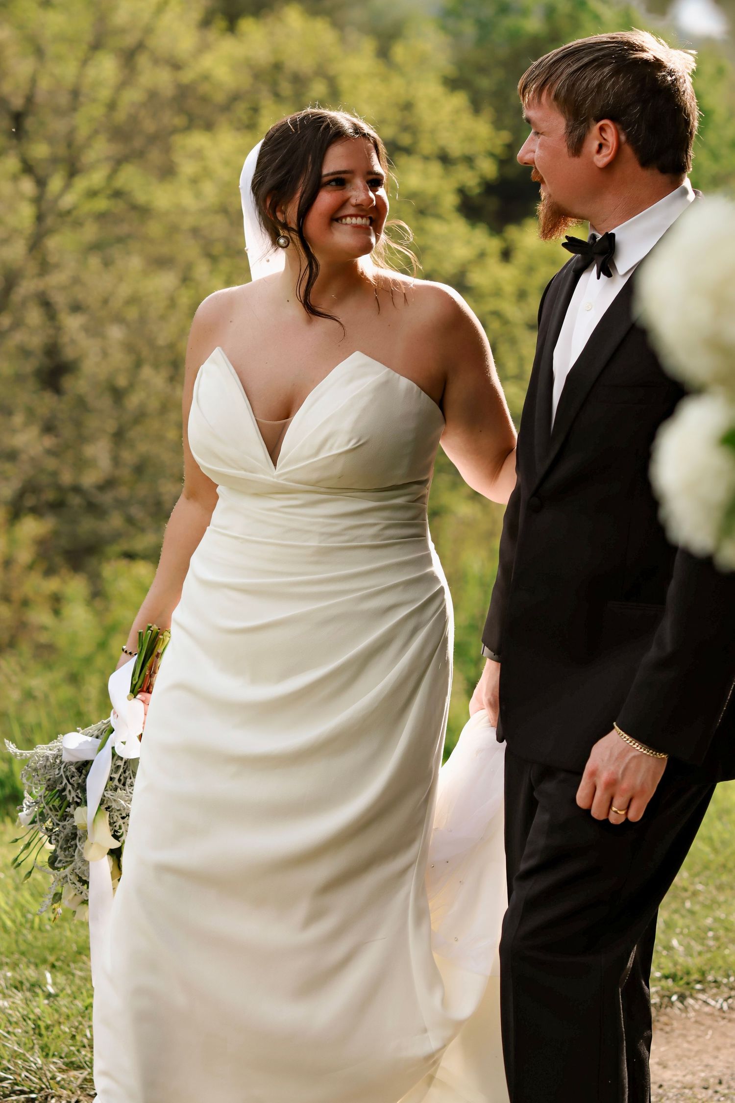 Bride in strapless white gown smiles at groom in black tuxedo outdoors with green trees in background.