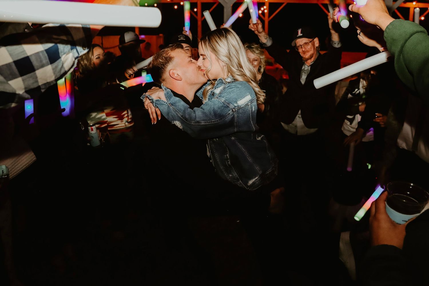 Couple sharing a romantic kiss at a vibrant party with colorful glow sticks and festive crowd.