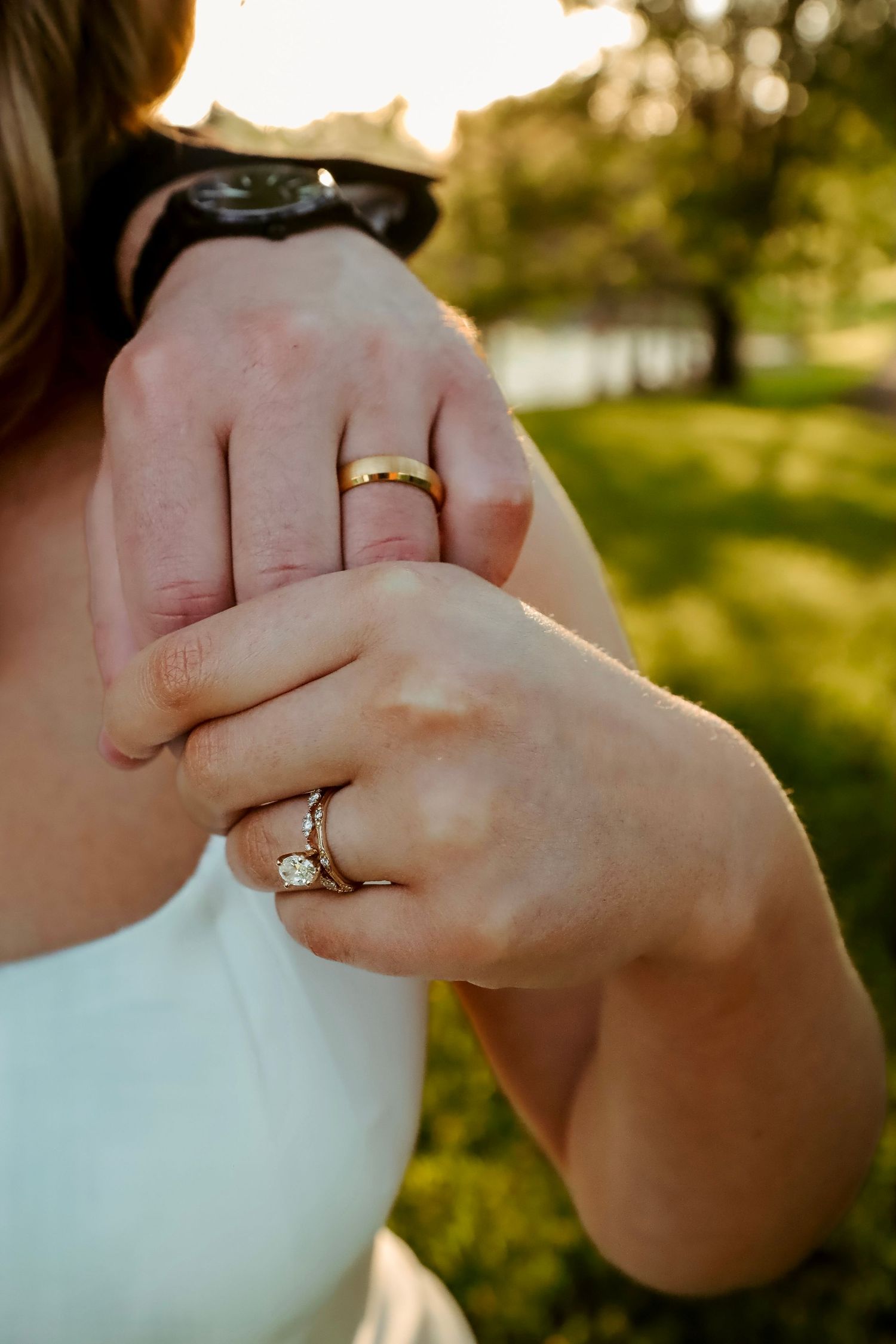 Close-up of couple's hands showing gold wedding band and diamond engagement ring outdoors in sunlight.