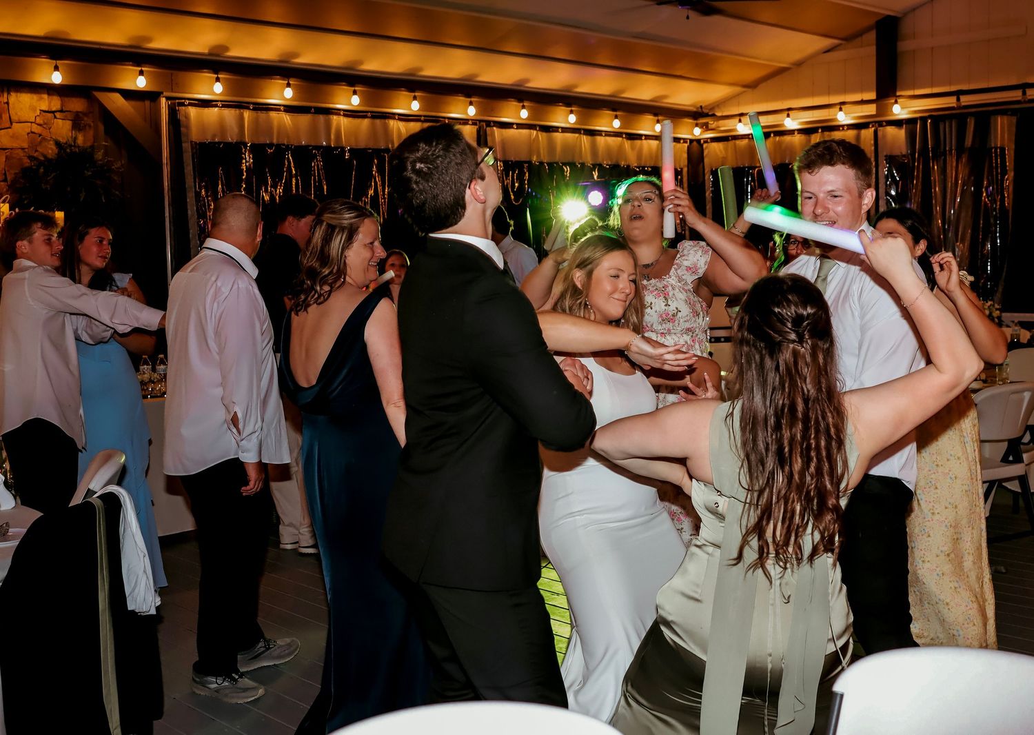 Wedding guests dancing energetically at a lively indoor reception party with string lights overhead.