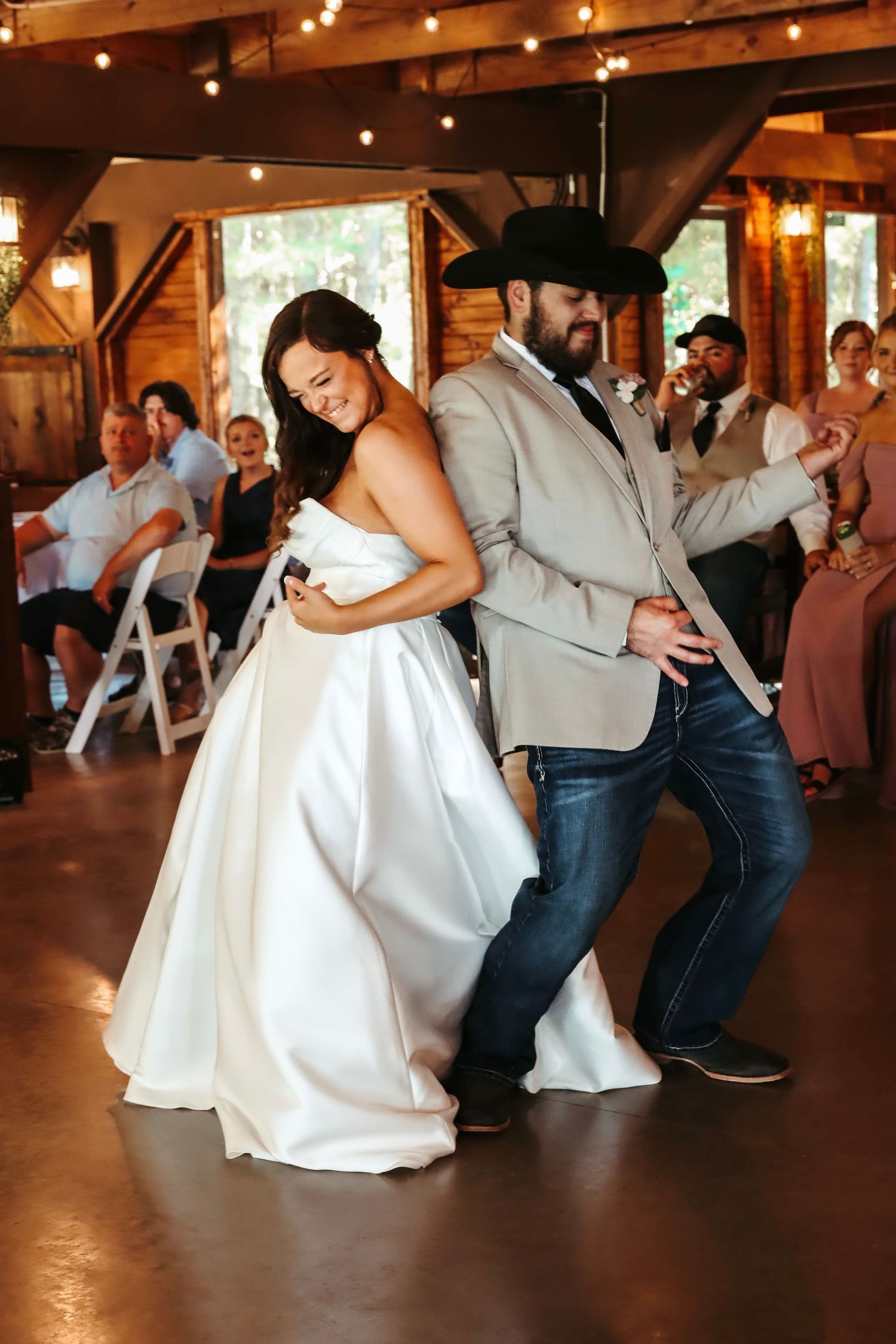 A bride and groom share a lively first dance at a rustic wedding reception with guests watching in the background.