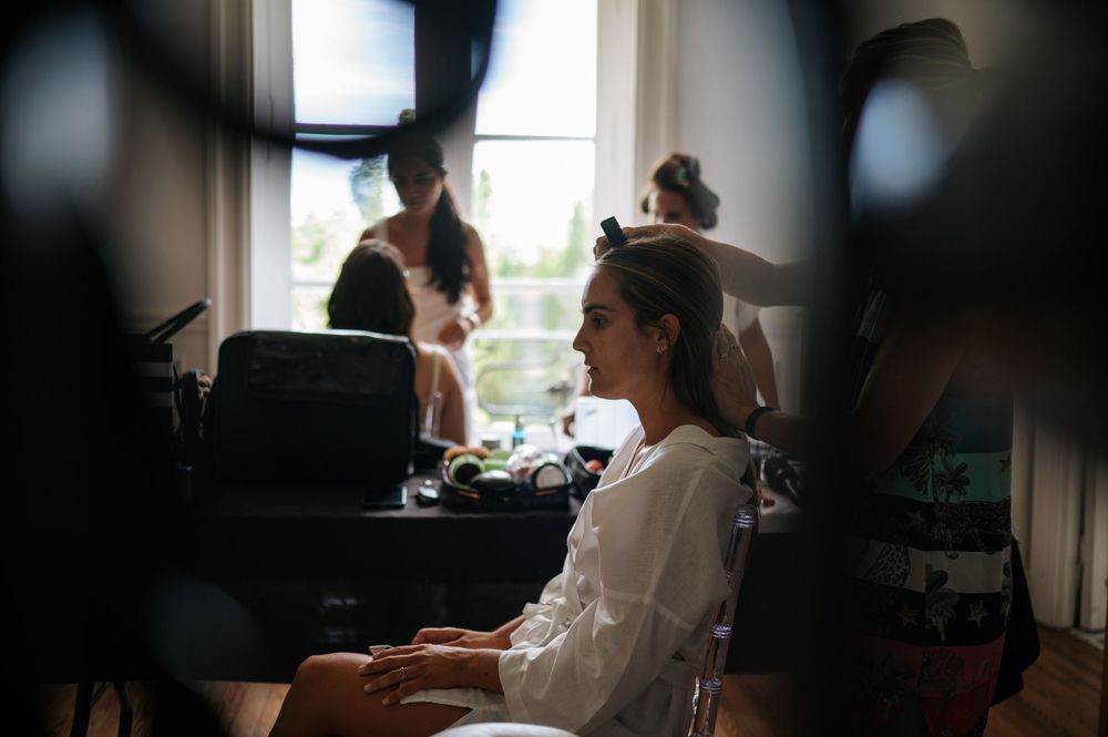 Bride in white robe smiling during hair and makeup preparations by a window.