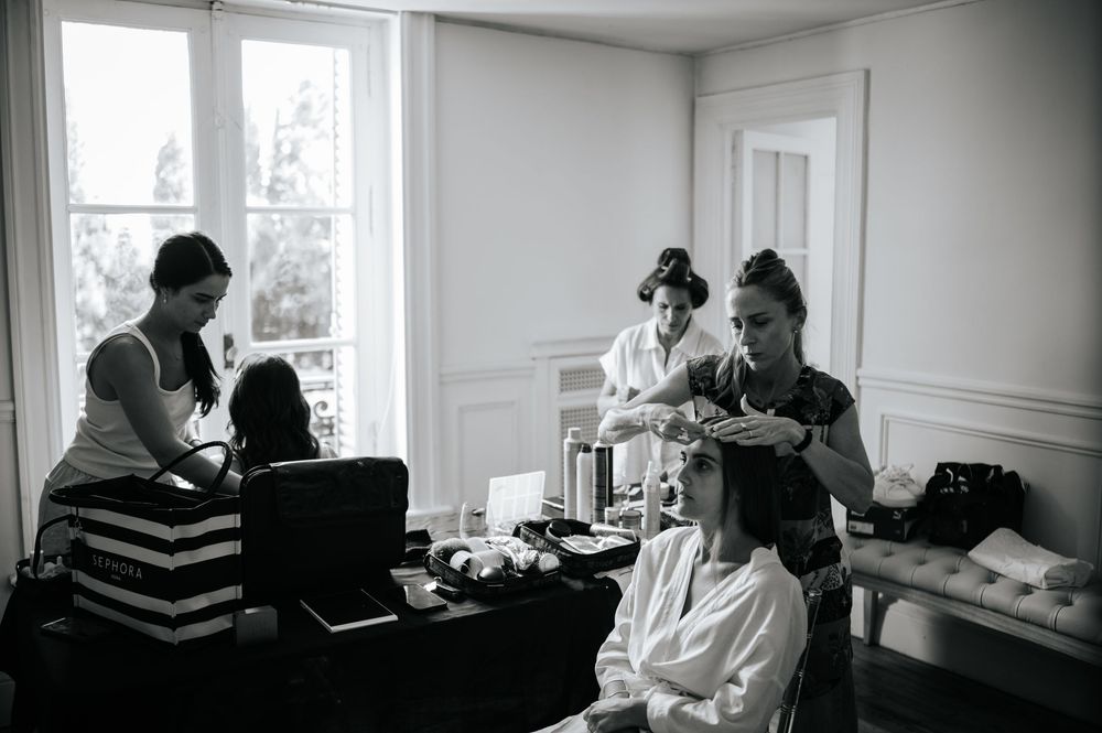 Black and white photo of bride getting hair styled by two stylists indoors.